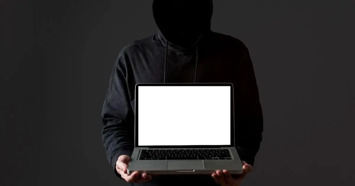 Indivíduo com capuz segurando um laptop com a tela em branco, contra um fundo escuro.