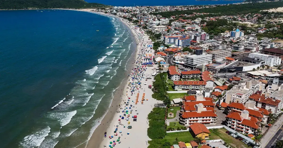 Praia com areia clara, ondas do mar, barracas, guarda-sóis coloridos e edifícios à beira-mar em Florianópolis.