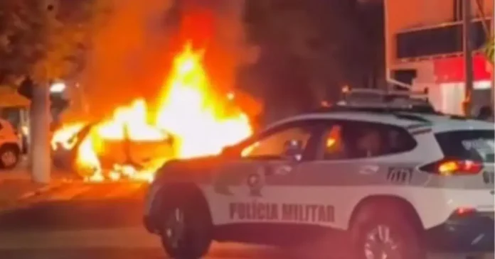 Veículo em chamas na rua, com um carro da polícia militar estacionado próximo.
