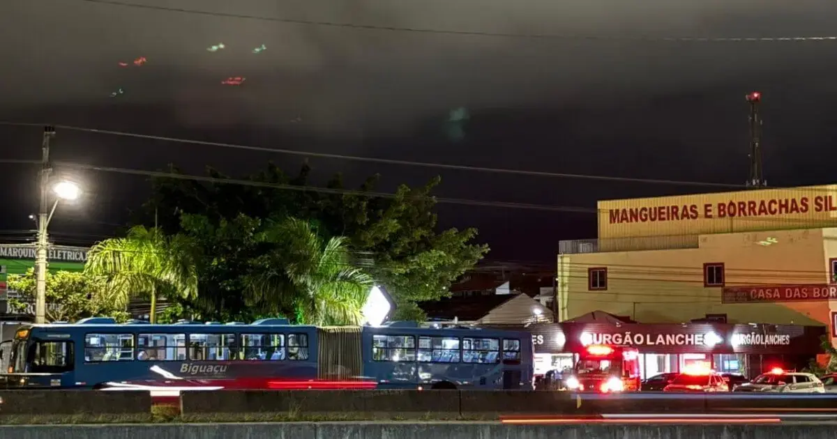 Homem esfaqueado dentro de ônibus na BR-101 em São José Ônibus azul de linha passando em frente a estabelecimentos comerciais iluminados à noite, sob céu nublado.
