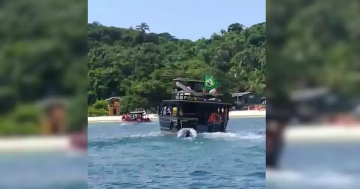 Homem morre após mal súbito na Ilha do Campeche Embarcação com bandeira do Brasil navegando em águas claras, com vegetação ao fundo e pessoas na praia.