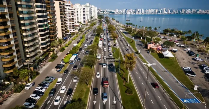 Vista de uma avenida com tráfego de veículos, cercada por prédios e com o mar ao fundo.