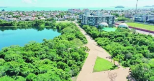 Vista aérea de um lago cercado por vegetação, com edifícios ao fundo e espaço para caminhadas.