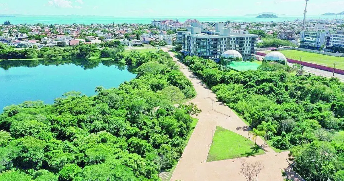 Inauguração do Parque Péricles Druck marca os 45 anos de Jurerê Internacional Vista aérea de um lago cercado por vegetação, com edifícios ao fundo e espaço para caminhadas.