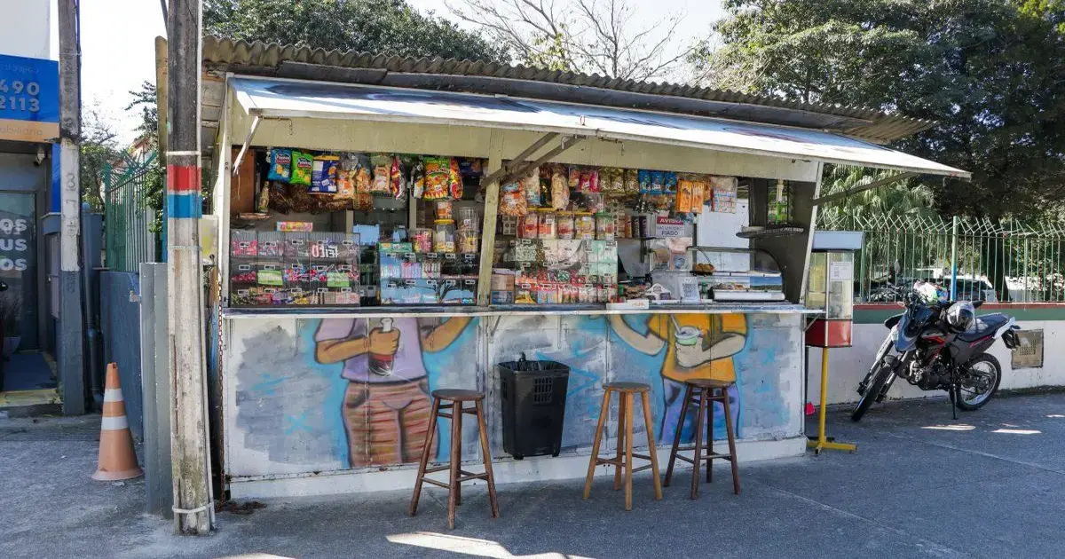 Quiosque com variedade de produtos à venda, incluindo lanches e bebidas, com dois bancos e uma moto estacionada ao lado.