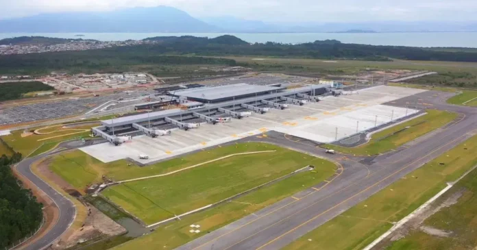 Vista aérea de um terminal de aeroportos com área de estacionamento e estrada ao redor, cercada por vegetação e montanhas ao fundo.