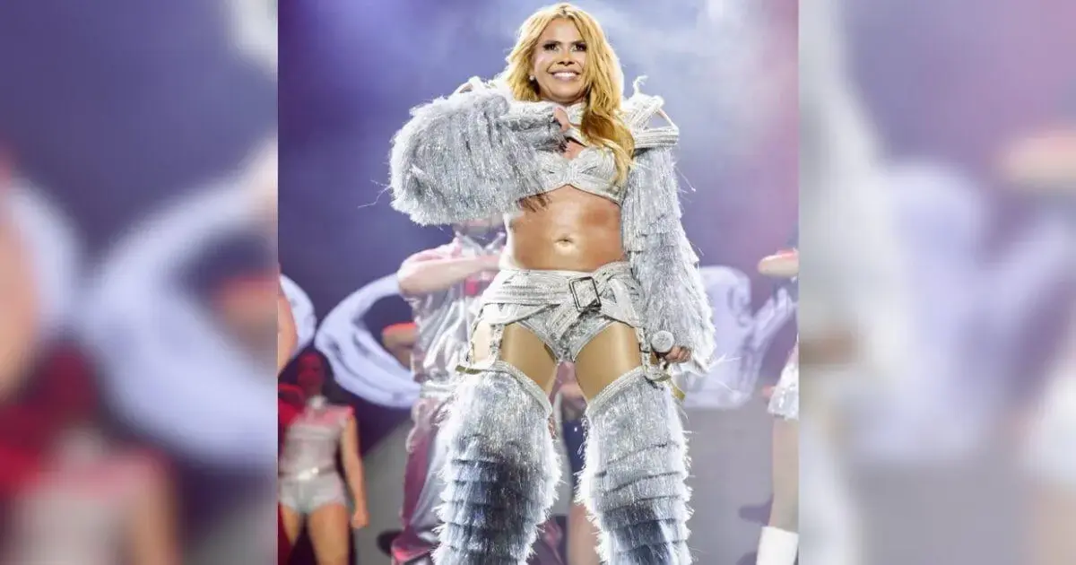 Joelma faz grande festa em Florianópolis durante Maratona Cultural Artista em um palco, vestindo traje prateado e dançando, com outros dançarinos ao fundo.