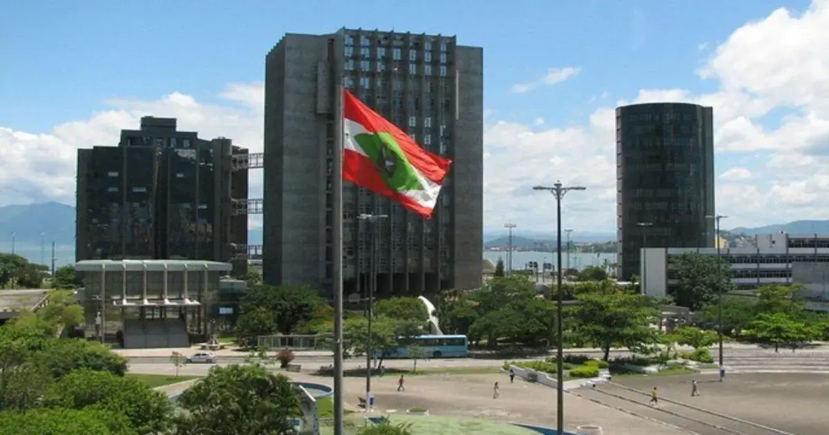 Vista de um espaço urbano com edifícios altos, uma bandeira, vegetação e um céu claro.