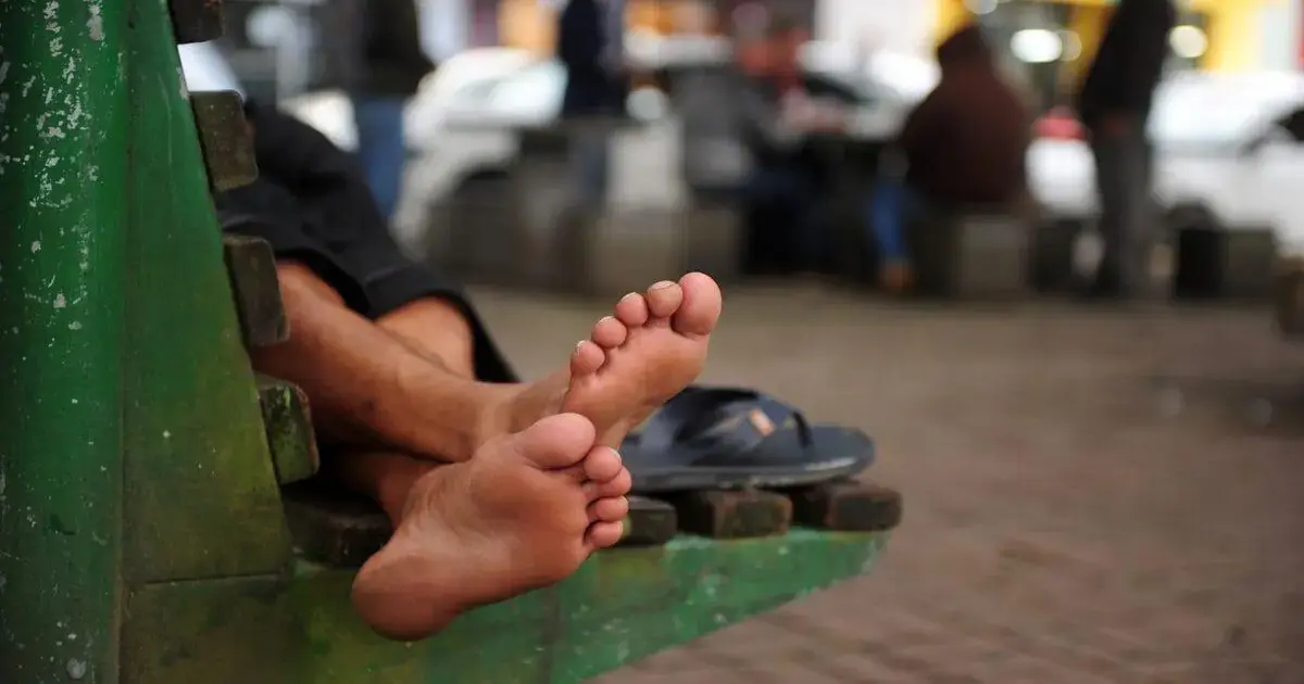 Pés descalços descansando sobre um banco em uma área pública, com pessoas ao fundo.
