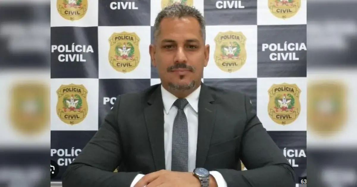 Retrato de um homem com terno, sorrindo e posando em frente a um fundo com logotipos da Polícia Civil.