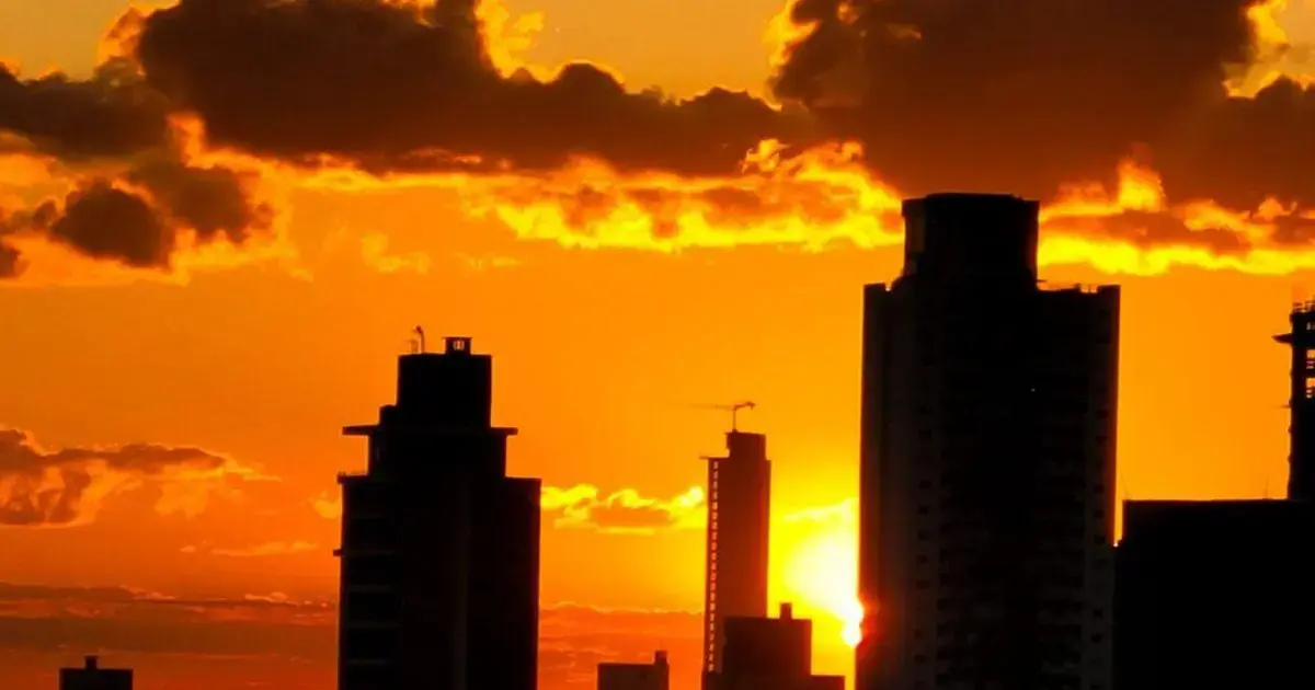 Silhuetas de prédios contra um pôr do sol alaranjado, com nuvens iluminadas no céu.