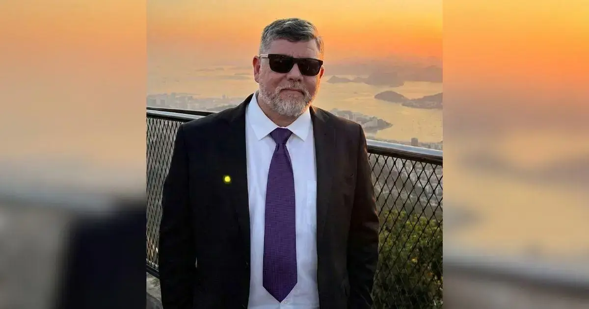 Homem com barba e cabelo curto usa terno escuro e óculos escuros, em frente a um pôr do sol.