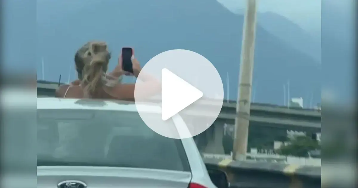 Mulher em pé no teto solar de um carro, segurando um celular, com vista para montanhas ao fundo.