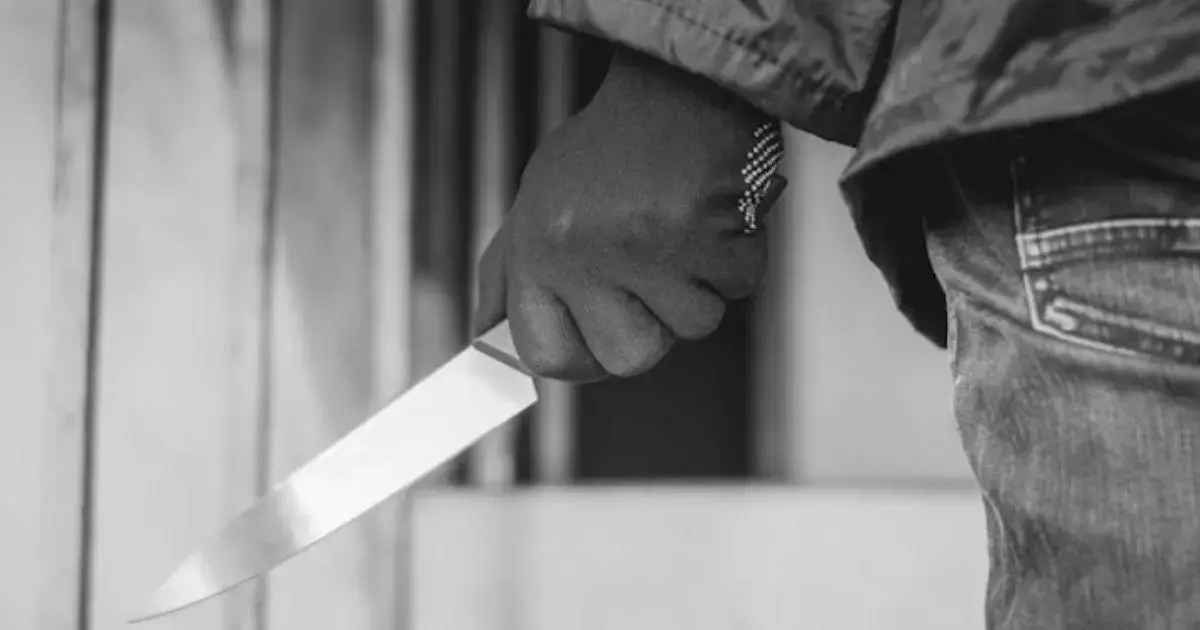 Mão segurando uma faca brilhante, com fundo desfocado mostrando uma entrada ou parede.