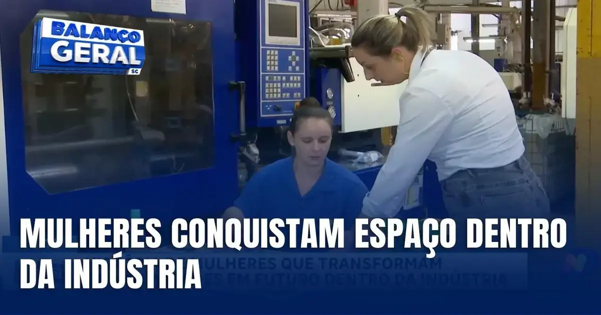 Duas trabalhadoras em ambiente industrial, com máquina ao fundo, discutindo sobre o trabalho realizado.