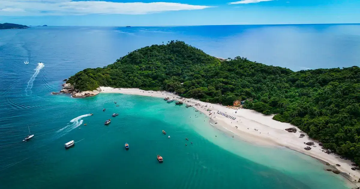Praia com águas claras e barcos ancorados, cercada por vegetação e uma pequena faixa de areia.