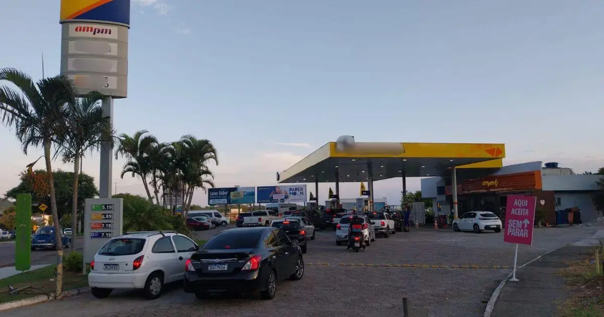 Posto de combustíveis com filas de veículos aguardando abastecimento, painel de preços visível.
