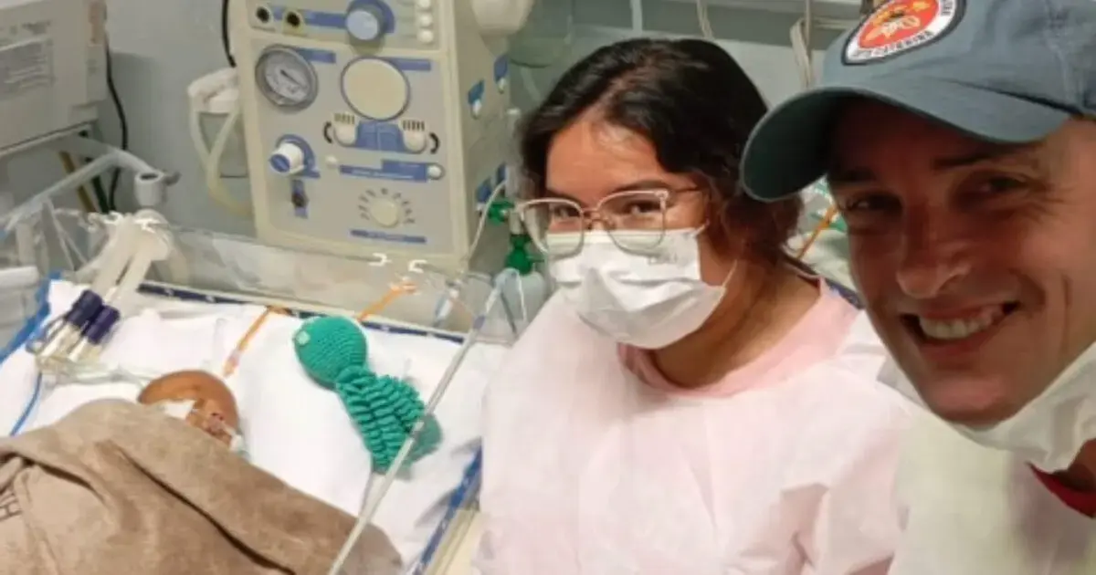 Mulher e homem sorrindo em um ambiente hospitalar, com um bebê em um berço ao fundo, cercado por equipamentos médicos.