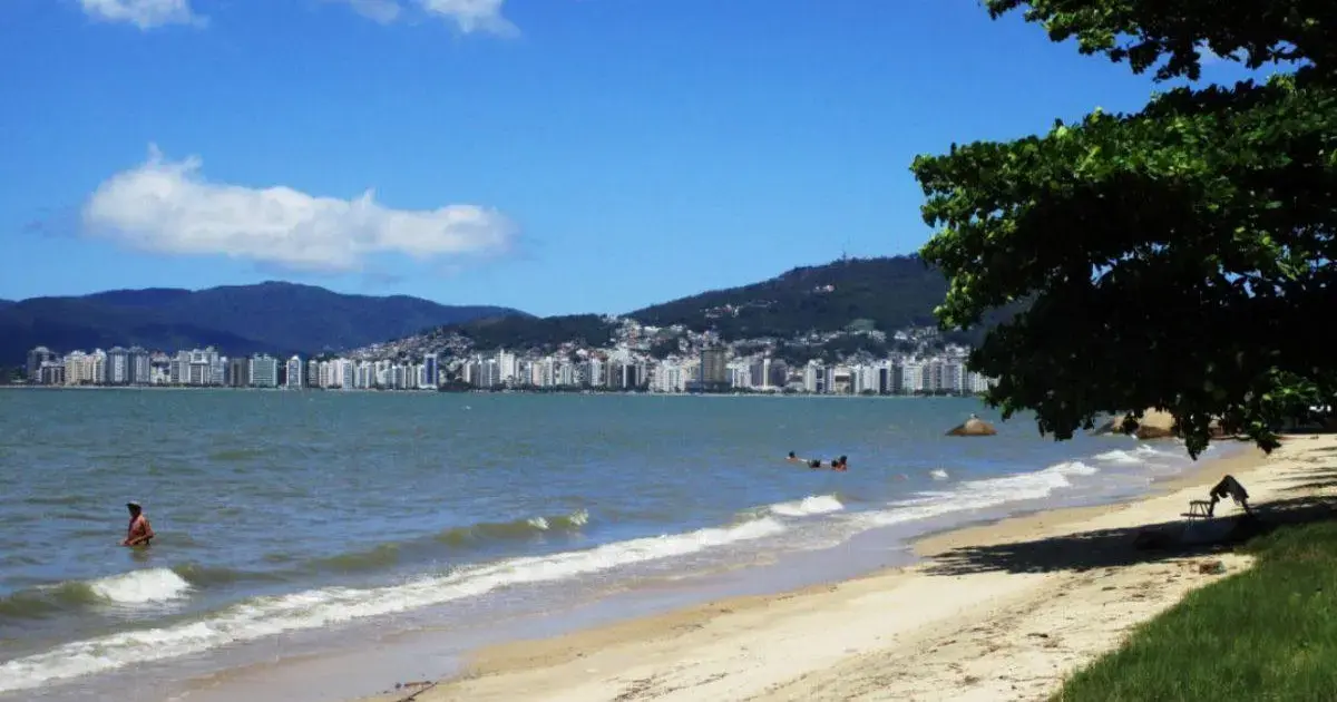 Vista da praia com água e pessoas nadando, com prédios ao fundo e montanhas.