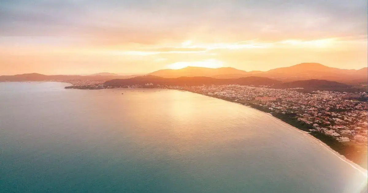Praias de Jurerê Próprias para Banho em 2026 Vista panorâmica de uma praia ao pôr do sol, refletindo tons laranja e dourado no mar calmo.