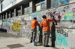 Duas pessoas em uniformes laranja limpam graffiti de uma parede com tinta e adesivos visíveis.
