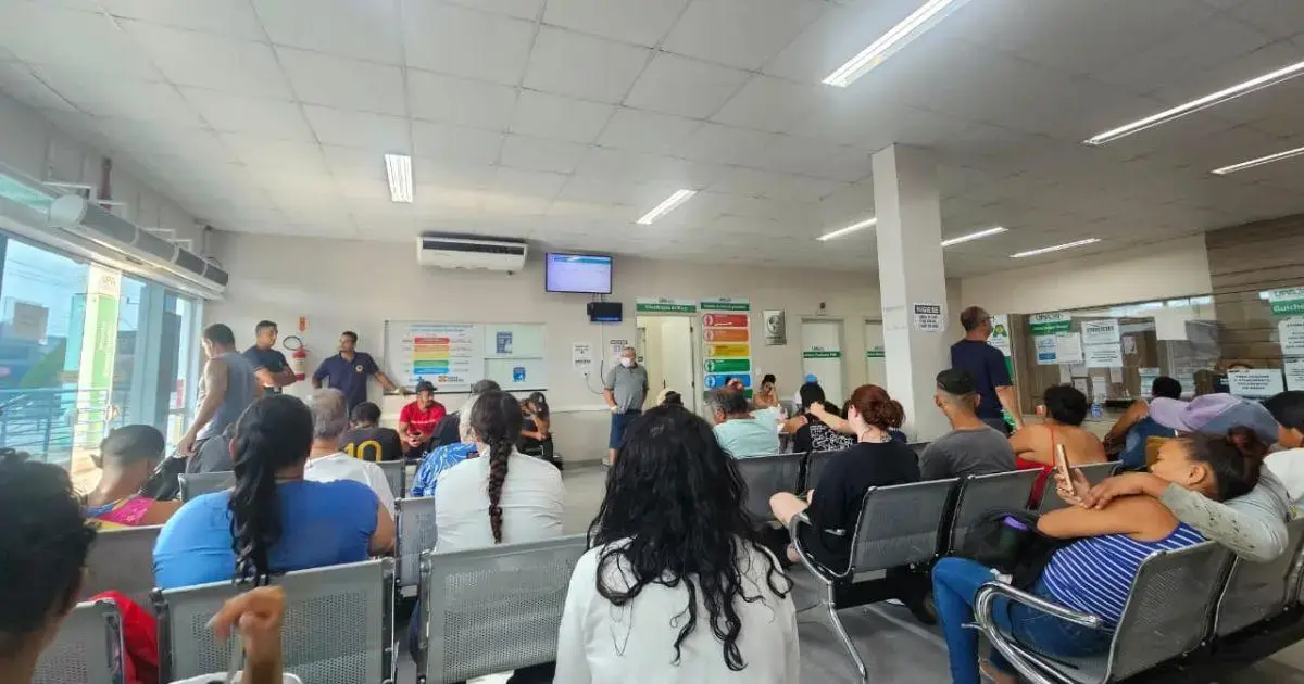 Sala de espera de uma unidade de saúde com pacientes sentados e funcionários conversando ao fundo.
