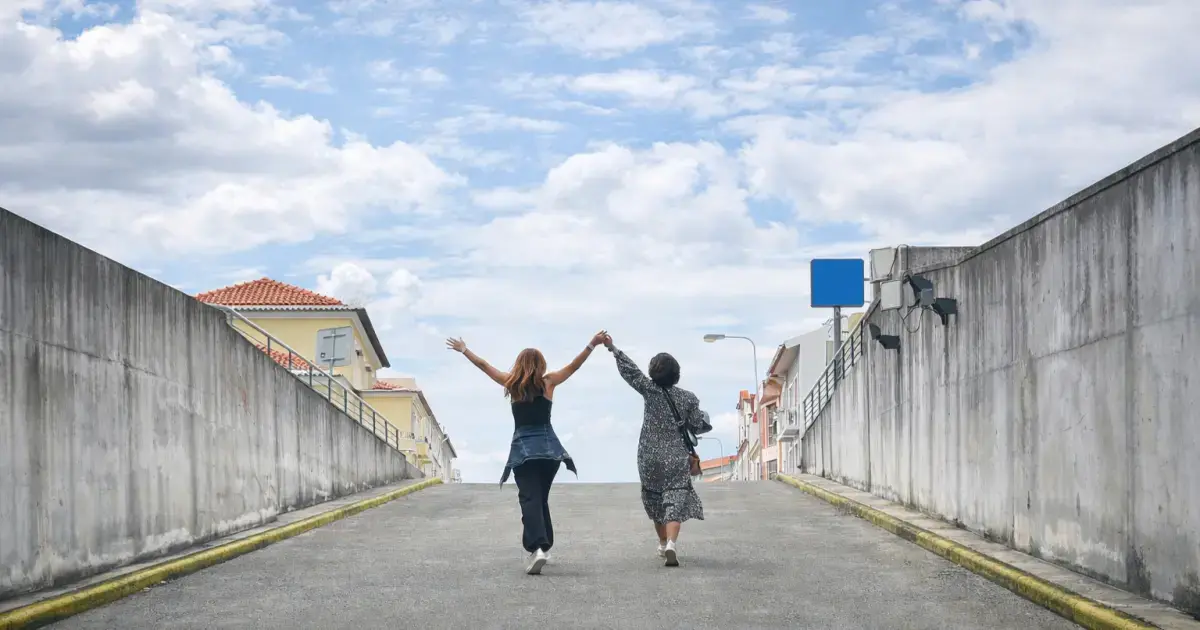 Duas mulheres caminham de mãos dadas por uma rua rodeada de casas sob um céu azul com nuvens.