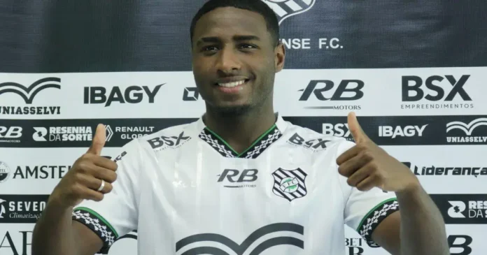 Atleta sorrindo e fazendo sinal de positivo, vestindo camisa branca com detalhes verdes e pretos, em fundo com logotipo do clube.