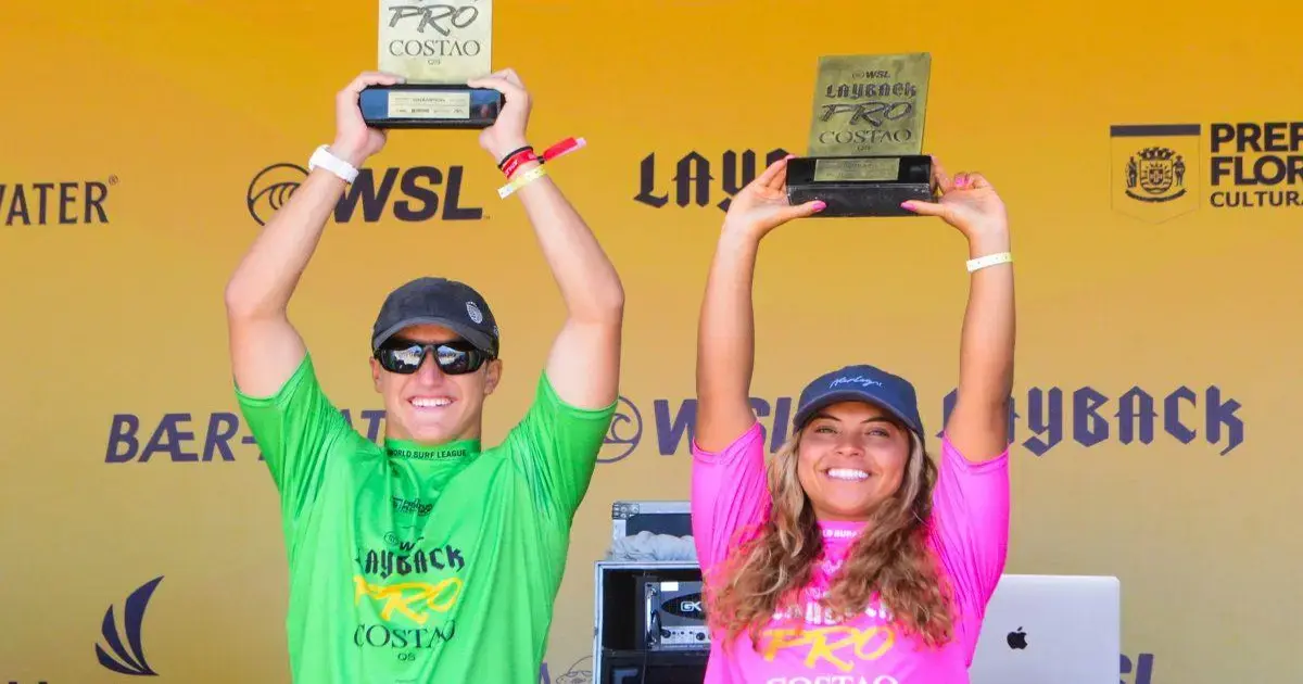 Duas pessoas levantam troféus em um evento de surfe, com fundo amarelo e logos da competição.