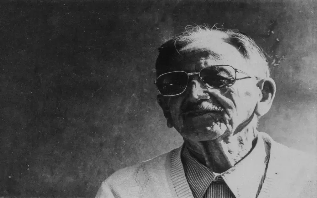 Retrato em preto e branco de um homem idoso usando óculos escuros e um suéter claro, com fundo escuro.