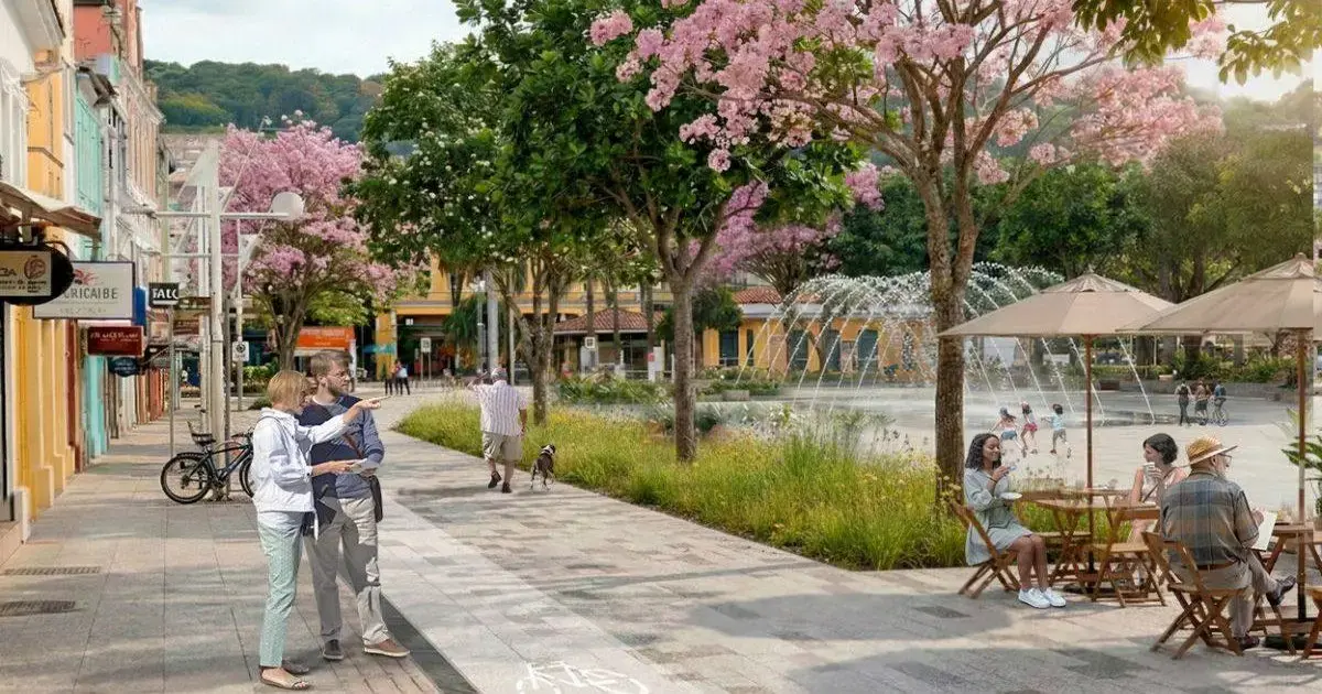 Caminho arborizado com flores, pessoas conversando, sentadas em mesas e uma fonte ao fundo.