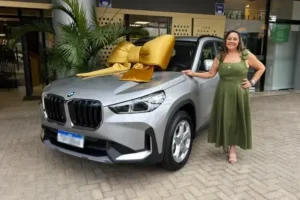 SUV prateado com laço dourado no capô, acompanhado por uma mulher vestindo um vestido verde em ambiente externo.