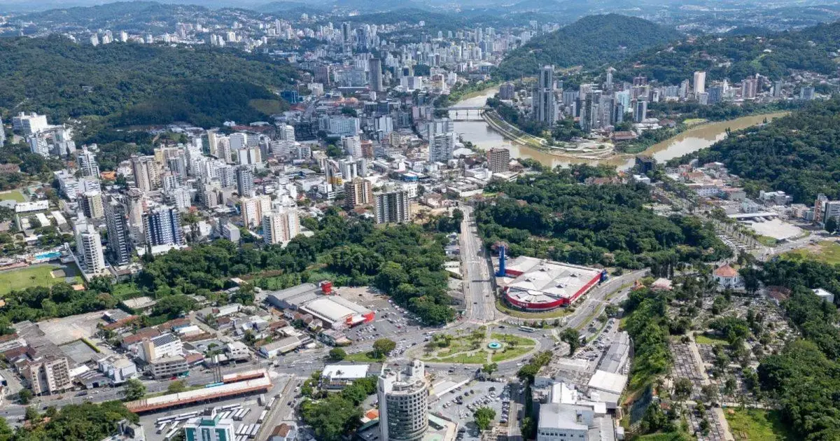 Vista aérea de uma cidade com prédios, áreas verdes, um rio e uma rodovia visível.
