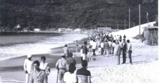 Pessoas caminhando pela praia, com mar e montanhas ao fundo, em uma cena de época.