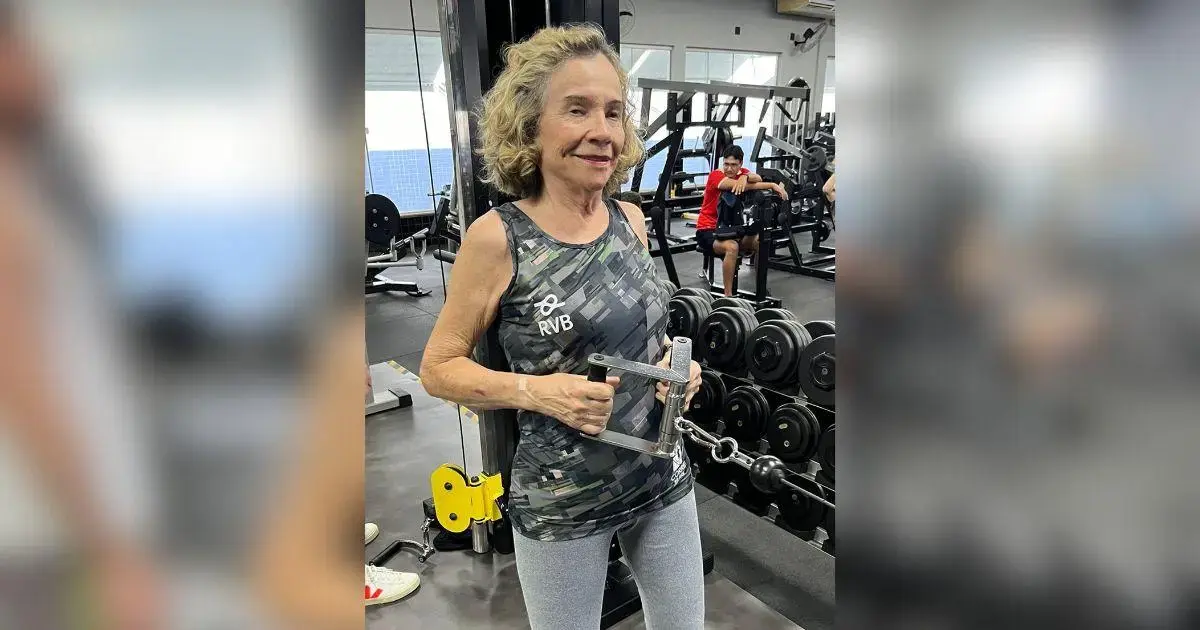 Vovó Maromba Inspira Mulheres em Santa Catarina Mulher de cabelo cacheado em academia segurando equipamento de musculação, com pesos ao fundo.