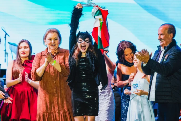 Criança vestida de super-heroína ergue um troféu enquanto segura uma bandeira, rodeada por aplausos e sorrisos.