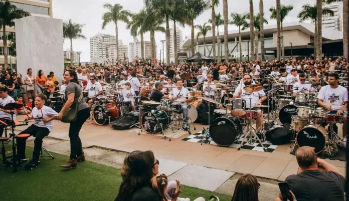 Um grande grupo de bateristas se apresenta em público, cercado por uma plateia animada e árvores.