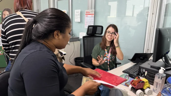 Duas pessoas interagindo em uma mesa, uma com documentos na mão e a outra sorrindo, enquanto um computador e brinquedo estão visíveis.