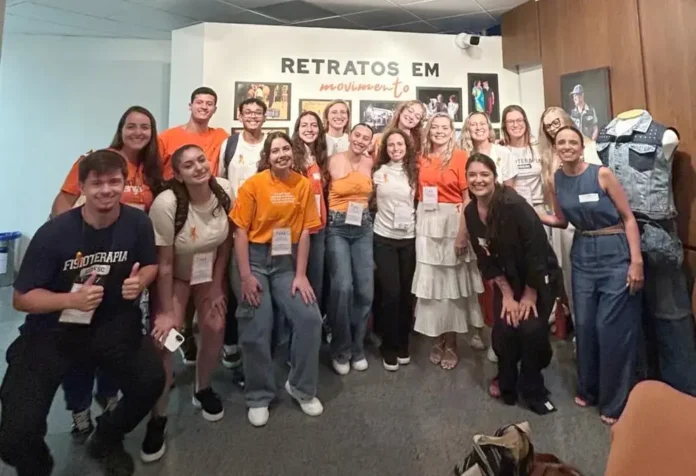 Grupo de pessoas sorrindo em um evento, com camisetas laranjas, em frente a uma parede com o texto "Retratos em movimento".