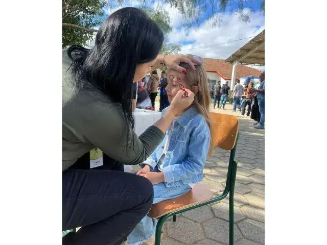 Uma criança sentada em uma cadeira enquanto uma pessoa a pinta, com outras pessoas ao fundo.