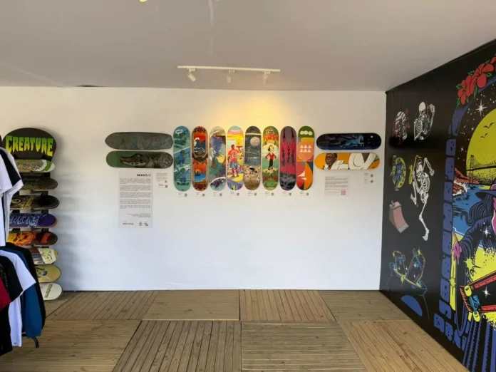Espaço de arte com pranchas de skate coloridas expostas na parede e roupas penduradas à esquerda.