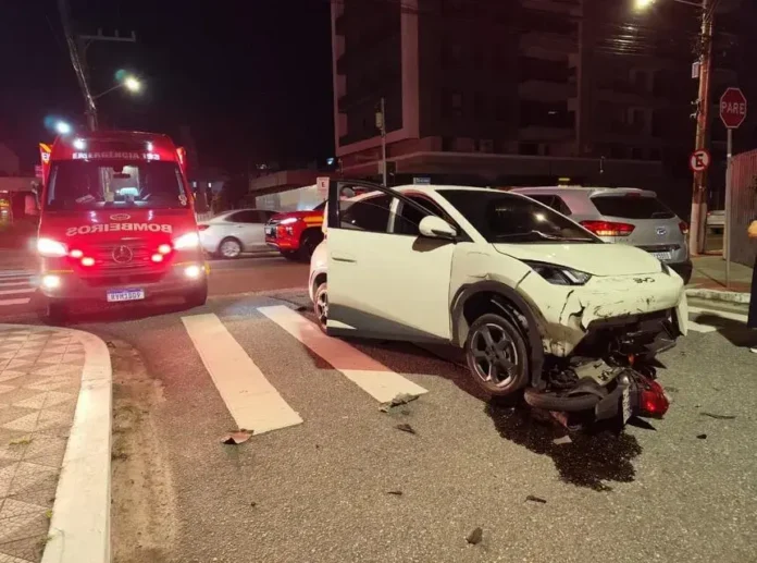 Veículo branco danificado em acidente, com a porta aberta, próximo a uma ambulância dos bombeiros.