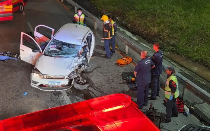 Veículo acidentado na rodovia com equipes de resgate e ambulância ao fundo.