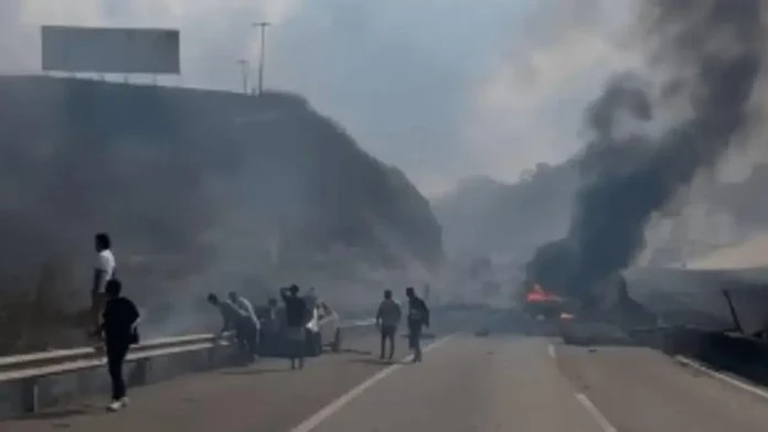 Caminho com fumaça e fogo, pessoas ao lado de veículos danificados e destroços na pista.