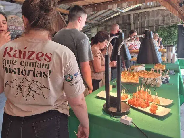 Grupo de pessoas em um evento, com mesa de petiscos e bandejas em um ambiente rústico e ao ar livre.