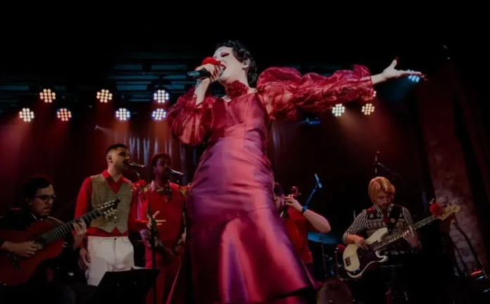 Artista em performance vestida com traje vermelho, cantando em palco com músicos e instrumentos ao fundo.