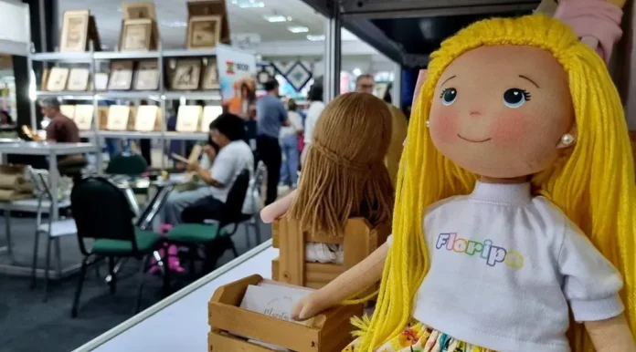 Boneca com cabelos longos e camiseta escrita "Floripa", ao fundo, visitantes em feira de artesanato.