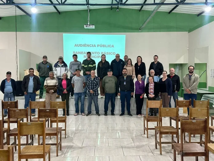 Grupo de pessoas em uma audiência pública sobre saneamento básico, com cadeiras vazias ao fundo e projeção no painel.