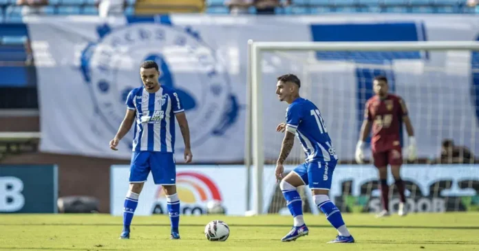 Dois jogadores em campo, um pronto para executar uma cobrança e o outro observando, com bandeira de time ao fundo.
