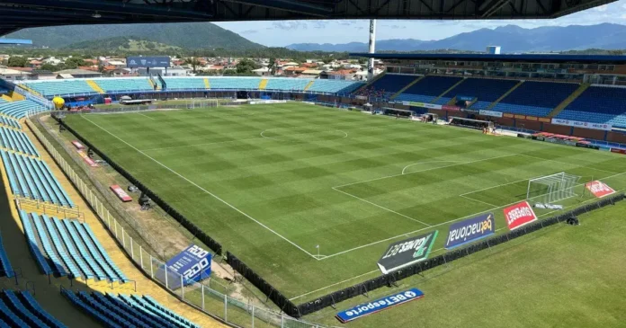 Campo de futebol em um estádio com arquibancadas azuis e gravato para o jogo, cercado por montanhas ao fundo.